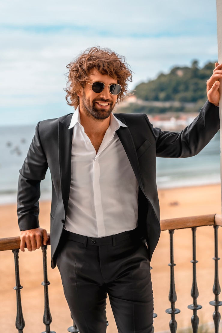 Foto estilo de rua com um jovem homem caucasiano de cabelos escuros em um terno preto e uma camisa branca em um hotel de luxo, com óculos de sol, editorial de moda. encostado na janela sério com o mar atrás