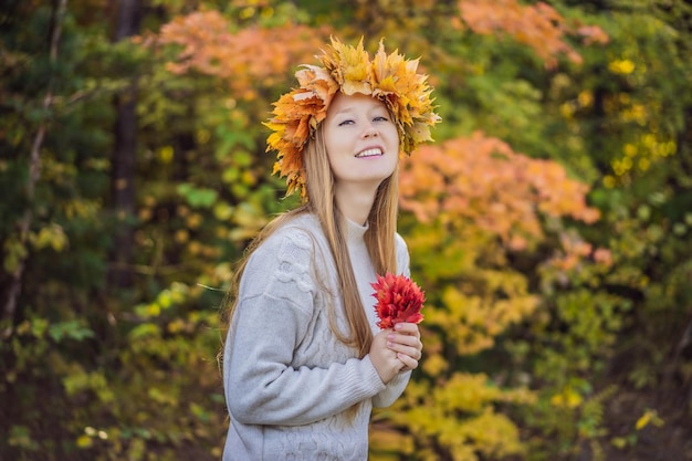 Estilo de vida ao ar livre retrato próximo de uma jovem loira encantadora vestindo uma coroa de folhas de outono Sorrindo caminhando no parque de outono Vestindo um elegante pulôver de malha Coroa de folhas de plátano