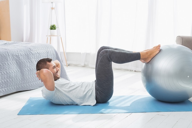 Estilo de vida ativo. Homem positivo e ativo colocando as pernas em uma bola médica enquanto se exercita em casa