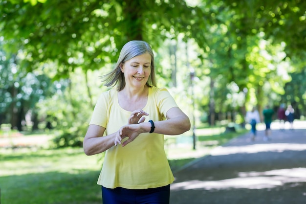 Estilo de vida ativo Mulher bonita sênior correndo no parque pela manhã Olha e monitora o pulso no relógio em sua mão Vai para esportes Sente bons sorrisos