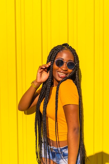 Estilo de vida, atraente garota negra com longas tranças, camisetas amarelas e jeans curtos em uma parede amarela. Com óculos escuros na sessão urbana na cidade, dançarina armadilha, mandando beijos no ar