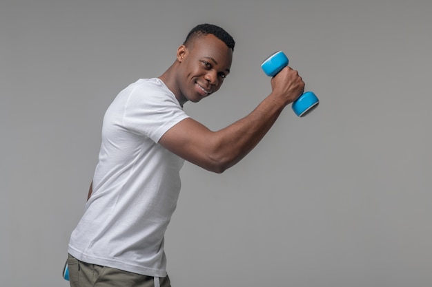 Estilo de vida. Atraente homem musculoso e alegre com halteres azul claro praticando esportes no estúdio