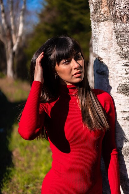 Estilo de vida da primavera. Retrato de uma jovem morena em um parque com um vestido vermelho descansando e curtindo a natureza ao lado de uma árvore