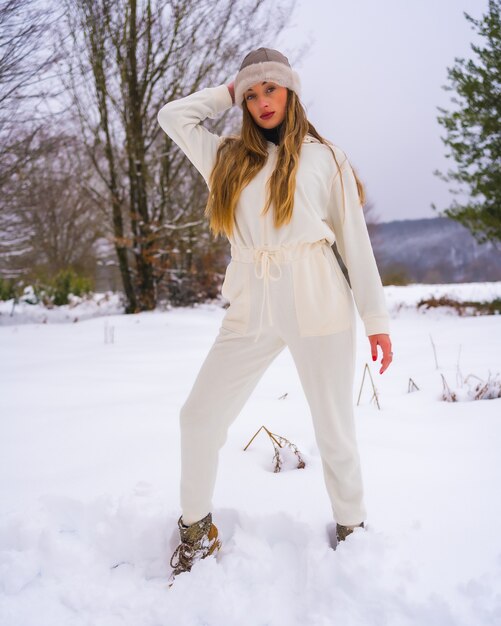 Estilo de vida de inverno, jovem loira caucasiana com roupa de inverno branca e chapéu de lã na neve ao lado de pinheiros nevados, férias na natureza