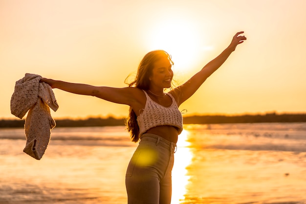 Foto estilo de vida de verão. uma jovem mulher caucasiana loira com um suéter branco curto de lã em um pôr do sol na praia. aproveitando a tarde de verão