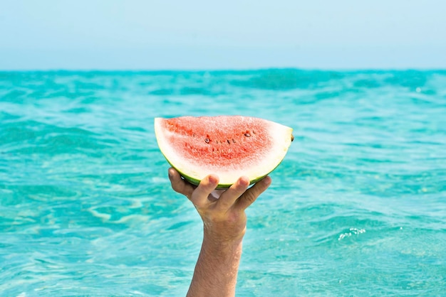 Estilo de vida do conceito de horário de verão Uma mão segurando um pedaço de melancia vermelha fresca contra um fundo de água do mar oceano azul Comida saudável e pessoas de férias Vitamina fruta Estilo de vida tropical feliz