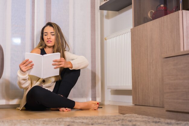 Foto estilo de vida em casa, jovem loira caucasiana com um café sorrindo, lendo um livro na sala de estar, sentada no chão