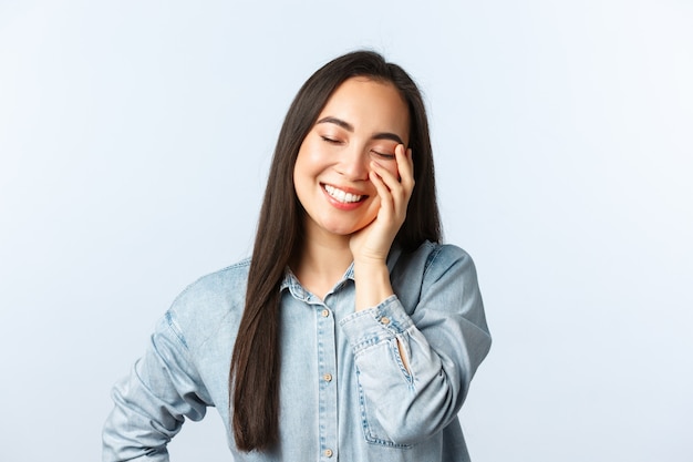 Estilo de vida, emoções de pessoas e conceito de beleza. Uma linda garota asiática feliz sonhadora rindo, corando e sorrindo com os olhos fechados, tocando a pele limpa, fundo branco