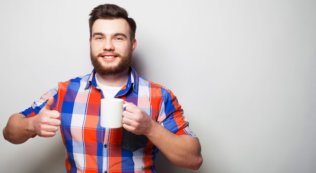 Foto estilo de vida, felicidade e conceito de pessoas: jovem barbudo com uma xícara de café na mão e mostrando ok, contra um fundo cinza.