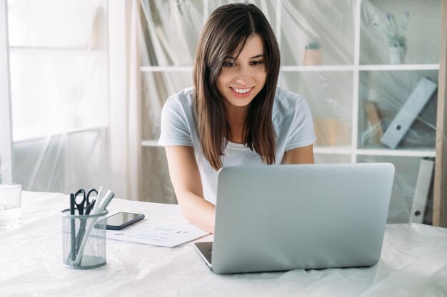 Estilo de vida freelancer Realocação em casa Trabalho remoto Escritório virtual Mulher feliz e satisfeita trabalhando on-line usando laptop em nova sala de luz moderna com móveis cobertos