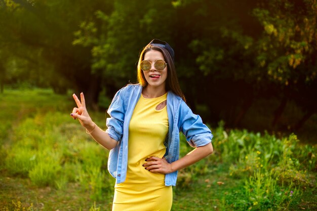Estilo de vida, garota feliz em óculos de sol e um boné em roupas brilhantes em um parque ao pôr do sol