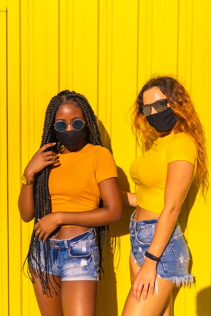 Estilo de vida, garota negra e garota loira caucasiana em camisetas amarelas em uma parede amarela. muito feliz e sorridente com máscaras na pandemia de coronavírus, novo normal, covid-19