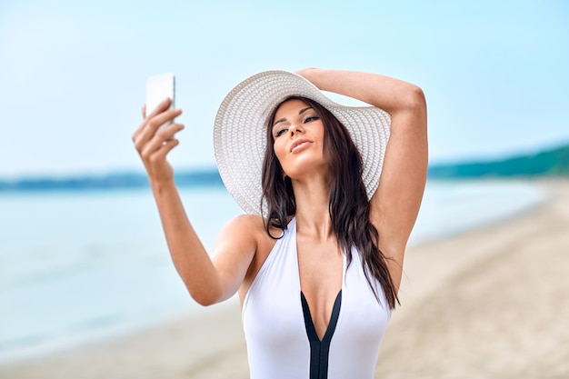 estilo de vida, lazer, verão, tecnologia e conceito de pessoas - jovem sorridente ou adolescente de chapéu de sol tomando selfie com smartphone na praia
