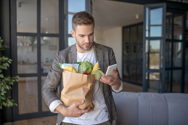 Estilo de vida moderno. Um homem segurando um pacote de vegetais e usando seu smartphone