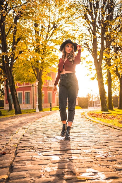 Estilo de vida outono ao pôr do sol loira Mulher caucasiana com um suéter vermelho e chapéu preto curtindo a natureza em um parque com árvores Caminhando no belo caminho do parque da cidade