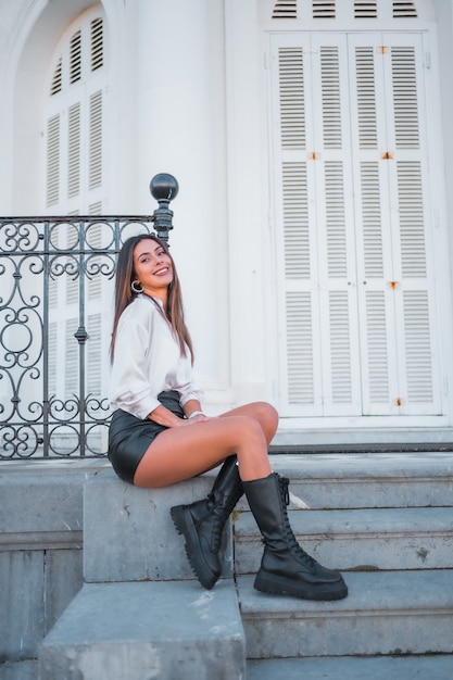 Estilo de vida, pose da moda de uma jovem morena de saia de couro preta e camisa sorrindo em uma escada em uma tarde de outono, foto vertical