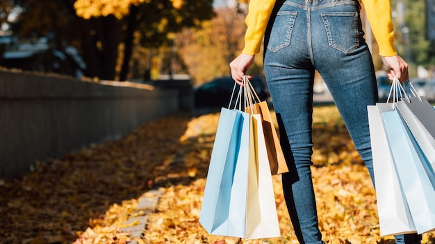 Estilo y moda de mujer Vista posterior recortada de dama en jeans de pie con bolsas de compras Desenfoque de fondo de la ciudad de otoño Espacio de copia