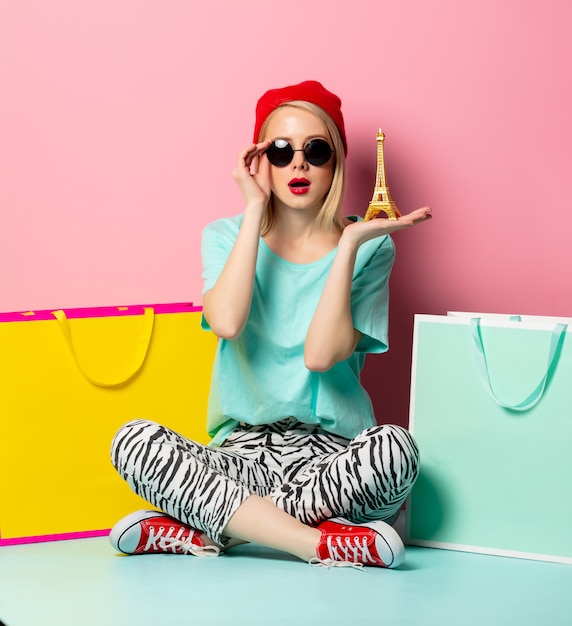 Foto estilo mujer en gafas de sol con bolsas de compras y pequeña torre eiffel en pared rosa