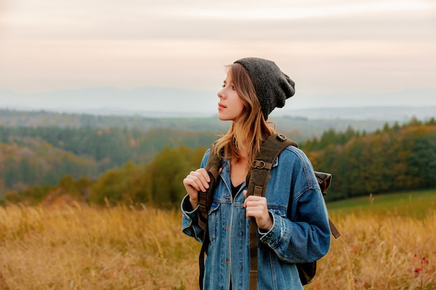 Estilo mulher de jaqueta jeans e chapéu com mochila na zona rural com montanhas