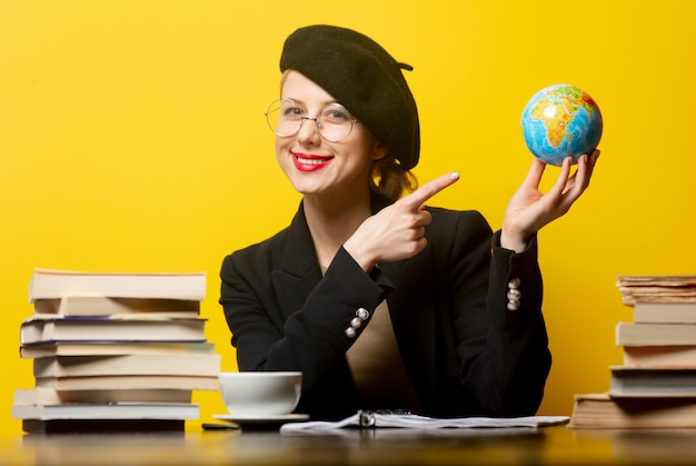 Estilo mulher loira na boina, sentado à mesa com livros ao redor e mantém o globo amarelo