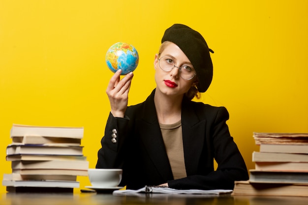 Estilo mulher loira na boina, sentado à mesa com livros ao redor e mantém o globo amarelo