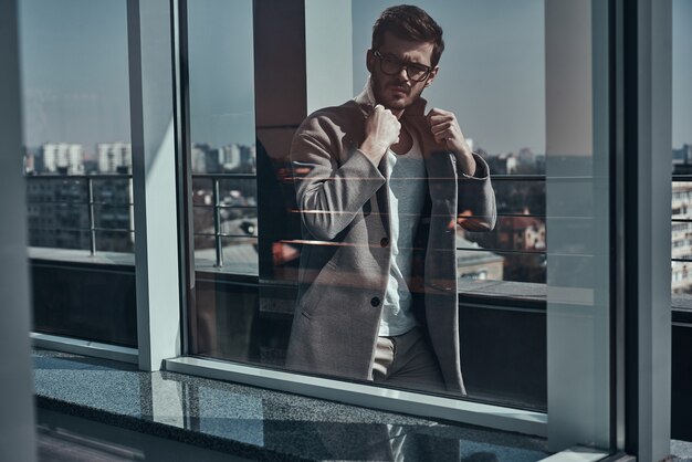 Estilo perfecto. A través de la ventana de un apuesto joven en gafas ajustando su chaqueta mientras camina al aire libre