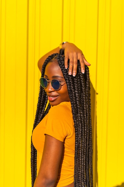 Estilo de vida, chica negra con largas trenzas, camisetas amarillas y jeans cortos en una pared amarilla. Pose de moda del atractivo con mirada seductora, fotografía callejera