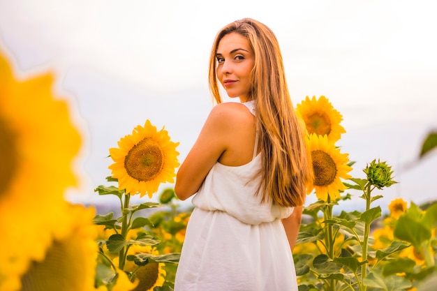 Estilo de vida, chica rubia entre girasoles con un vestido blanco y un cinturón mirando a la cámara