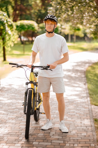 Estilo de vida. Chico guapo amable en camiseta y casco con pantalones cortos ligeros con bicicleta de pie en el parque el día de verano