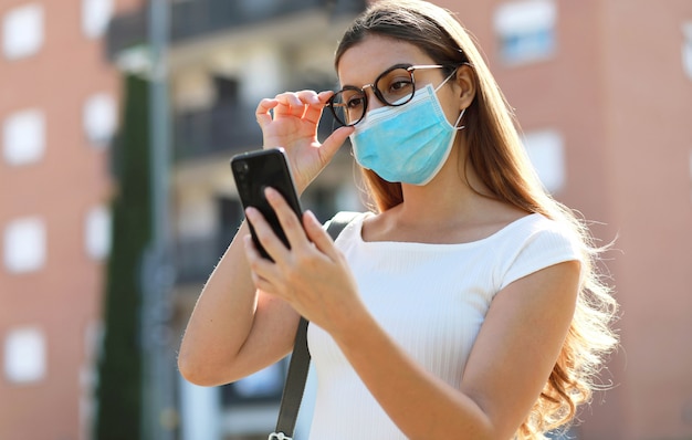 Estilo de vida de la ciudad chica hipster con estilo con máscara quirúrgica leyendo un mensaje en el teléfono móvil en la calle
