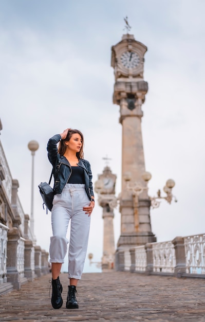 Estilo de vida en la ciudad con una chica rubia con pantalones blancos y una chaqueta de cuero. Caminando por una pendiente con relojes en el fondo