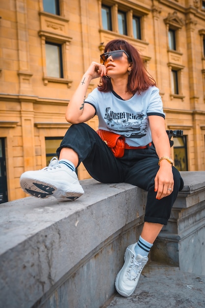 Estilo de vida en la ciudad. Una joven morena con gafas de sol, una camiseta blanca, una riñonera roja, pantalones negros y zapatillas blancas. Sentado y pensativo