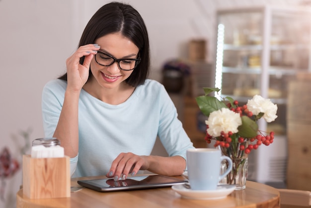 Estilo de vida conectado. Hermosa mujer alegre inspirada sonriendo y usando su gadget mientras se relaja en una cafetería.