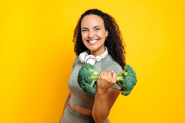 Foto estilo de vida deportivo motivado emocionado feliz de raza mixta mujer joven deportiva con auriculares en los hombros sosteniendo un kettlebell de brócoli imaginario de pie en un fondo amarillo aislado sonriendo haciendo ejercicio