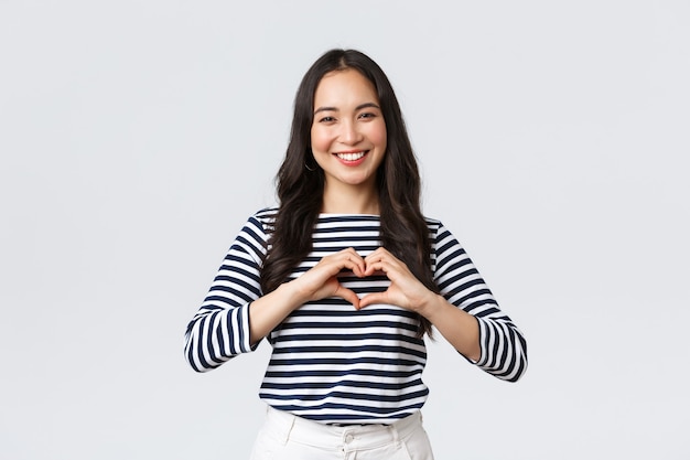 Estilo de vida, emociones de la gente y concepto casual. Hermosa mujer asiática adorable sonriente que muestra el signo del corazón y sonriendo, expresa simpatía o cuidado, de pie fondo blanco.