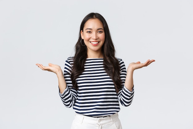 Estilo de vida, emociones de la gente y concepto casual. Linda mujer asiática sonriente presenta dos productos, se toma de las manos hacia los lados como si estuviera demostrando productos en las palmas, de pie fondo blanco