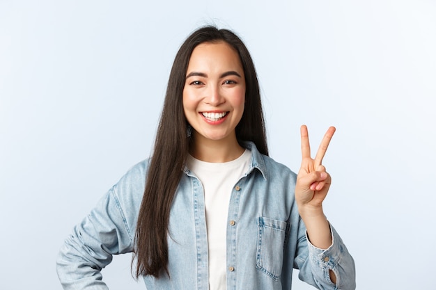 Estilo de vida, emociones de las personas y concepto de belleza. Alegre linda chica asiática, estudiante en la universidad que parece feliz, muestra el signo de la paz y una amplia sonrisa, de pie con fondo blanco.