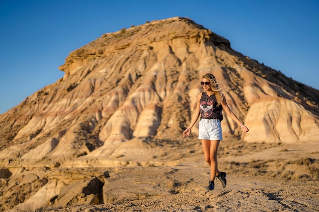 Estilo de vida, feliz chica caucásica rubia con botas negras, gafas de sol y pantalones cortos paseando por el desierto de Bardenas. España