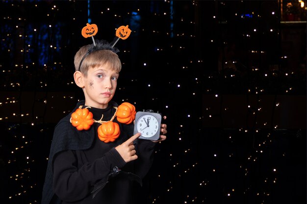 Estilo de vida de Halloween. Niño con una araña en la mejilla y perlas de calabaza sosteniendo un despertador negro sobre un fondo negro con bokeh. Es hora de celebrar Halloween. Feliz concepto de Halloween