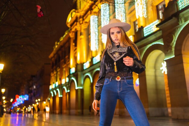 Estilo de vida de invierno, chica rubia caucásica con jeans y una chaqueta negra en la ciudad iluminada para Navidad por la noche