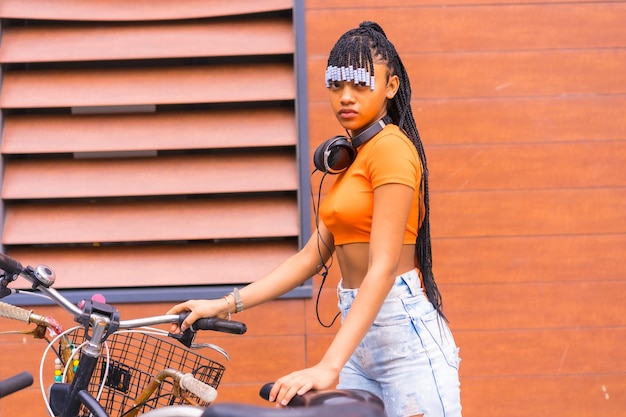 Estilo de vida con una joven bailarina de trap en la ciudad. Chica negra grind de etnia africana con camisa naranja en la ciudad. Cofing la bicicleta estacionada en la ciudad.