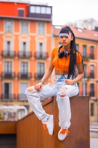 Foto estilo de vida con una joven bailarina de trap con trenzas. chica negra de etnia africana con camiseta naranja y pantalón vaquero. con fondo de casas naranja