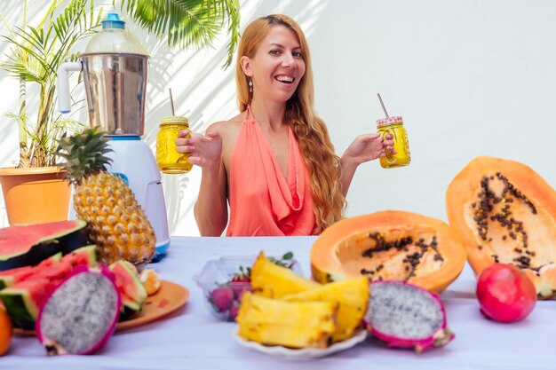 Estilo de vida mujer rubia mezclando frascos de vidrio de jugo de licuadora con batidos y paja en el balcón de verano.Frutas exóticas tropicales sobre la mesa.La limpieza del cuerpo de toxinas idea.temporada de verano