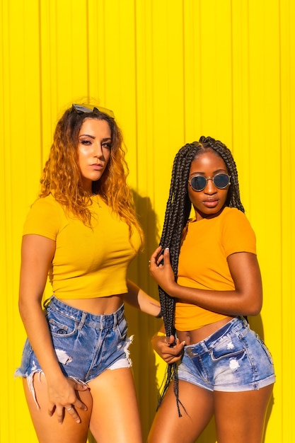 Estilo de vida de novias, chica negra con largas trenzas y rubia caucásica con camisas amarillas y jeans cortos en una pared amarilla. Sesión urbana con colores cálidos en la ciudad