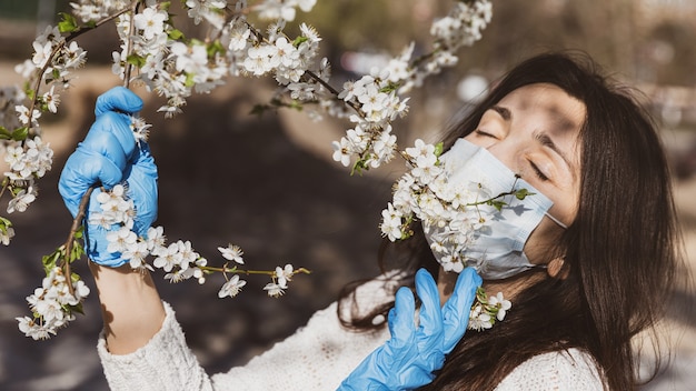 Foto estilo de vida en el nuevo concepto de entorno. primavera y la epidemia de coronavirus. una mujer joven con guantes médicos y una máscara protectora disfruta de árboles con flores de primavera