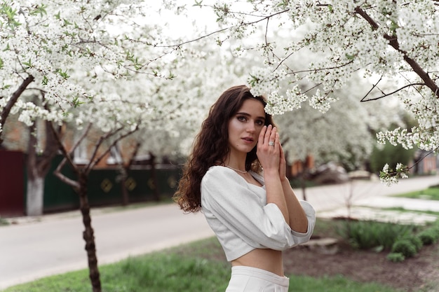 Foto estilo de vida primaveral. modelo posando cerca de árboles florecientes blancos sin campo al aire libre de máscara. chica soñando con el pelo rizado.