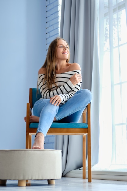 Estilo de vida retrato de cuerpo entero de una niña sentada en un sillón acogedor junto a la ventana en casa