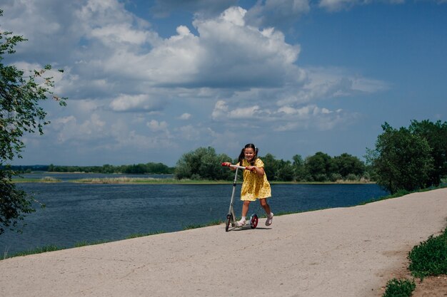 Estilo de vida retrato de cuerpo entero de un niño feliz riendo montando una scooter en un hermoso bosque a lo largo de ...