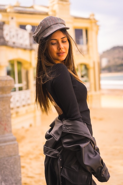 Estilo de vida, retrato de una joven morena caucásica sentada en unas escaleras con un vestido negro corto, con una boina y medias a cuadros junto a una playa, foto vertical