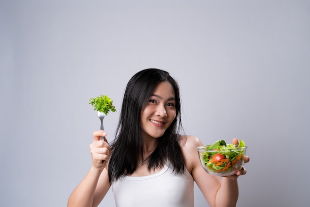 Estilo de vida saludable con concepto de comida limpia. Mujer asiática feliz comiendo ensalada aislada sobre la pared blanca.